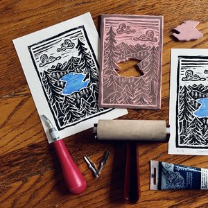 A linoleum cut stamp of a lake vista with two prints and block printing supplies
